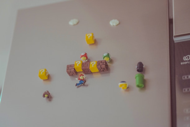 A collection of small, colorful Super Mario-themed magnets is attached to a refrigerator surface. These include Mario, question blocks, mushrooms, and a green pipe, resembling elements from the iconic video game series.