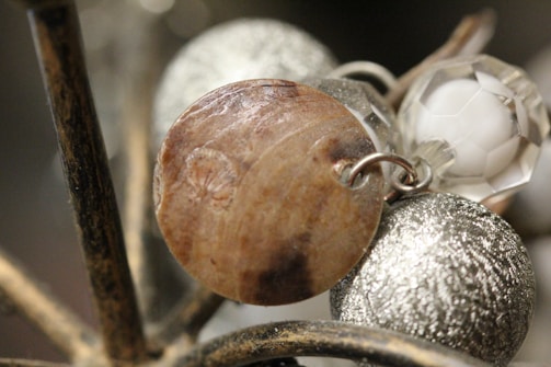 A close-up of a piece of jewelry with a wooden round element attached to a metal hoop, alongside a textured silver sphere and a faceted clear bead. The objects are intertwined on what seems to be a metallic structure.