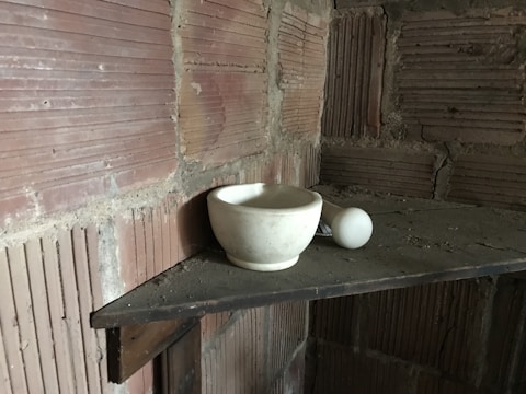 A white mortar and pestle sit on a dusty wooden shelf against a backdrop of exposed, textured brick walls.