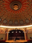 An ornate theater interior with an intricate, circular ceiling featuring elaborate carvings and designs in rich red and gold tones. Below the ceiling, a grand stage with a dark red curtain backdrop is illuminated by rows of bright lights. A person is seated at a grand piano on the stage, adding a sense of scale to the vast and opulent space. The sides of the stage are adorned with detailed murals depicting historical or classical scenes.