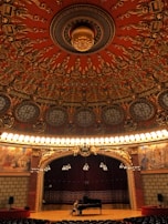 An ornate theater interior with an intricate, circular ceiling featuring elaborate carvings and designs in rich red and gold tones. Below the ceiling, a grand stage with a dark red curtain backdrop is illuminated by rows of bright lights. A person is seated at a grand piano on the stage, adding a sense of scale to the vast and opulent space. The sides of the stage are adorned with detailed murals depicting historical or classical scenes.