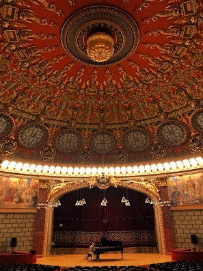 An ornate theater interior with an intricate, circular ceiling featuring elaborate carvings and designs in rich red and gold tones. Below the ceiling, a grand stage with a dark red curtain backdrop is illuminated by rows of bright lights. A person is seated at a grand piano on the stage, adding a sense of scale to the vast and opulent space. The sides of the stage are adorned with detailed murals depicting historical or classical scenes.