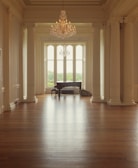 A spacious, elegantly decorated room featuring tall white columns and a glossy wooden floor. At the far end near tall windows with a view of greenery, there is a grand piano accompanied by a bench and a violin case. A chandelier hangs from the ceiling adding a touch of luxury to the setting.