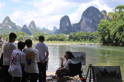 Several people are standing by a calm river, watching an artist painting landscapes. The setting includes a backdrop of lush greenery and dramatic karst mountains under a partly cloudy sky.