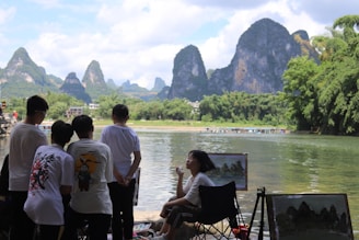 Several people are standing by a calm river, watching an artist painting landscapes. The setting includes a backdrop of lush greenery and dramatic karst mountains under a partly cloudy sky.