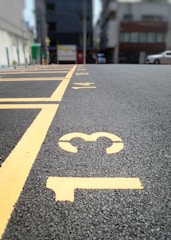 A close-up view of a paved parking lot with yellow painted lines and numbers on the ground. The numbers 13, 14, and 15 are clearly visible. In the background, there are buildings and cars, but they are out of focus.