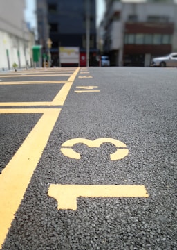 A close-up view of a paved parking lot with yellow painted lines and numbers on the ground. The numbers 13, 14, and 15 are clearly visible. In the background, there are buildings and cars, but they are out of focus.