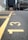 A close-up view of a paved parking lot with yellow painted lines and numbers on the ground. The numbers 13, 14, and 15 are clearly visible. In the background, there are buildings and cars, but they are out of focus.