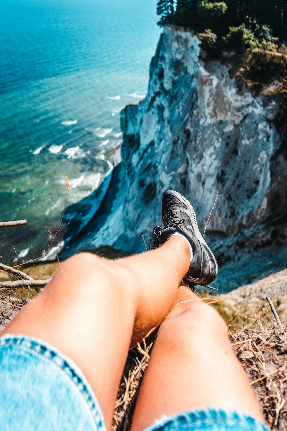 person wearing black-and-gray shoes and blue denim shorts sitting on mountain cliff