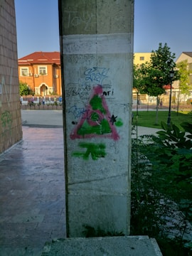 A pillar covered in various graffiti, including a large pink and green symbol resembling an anarchy sign. The background features a sidewalk, some trees, and a building with a red roof under a clear blue sky.