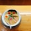 A bowl of ramen soup placed on a wooden surface. It contains various ingredients including two boiled eggs with partially soft yolks, a piece of seaweed, slices of pork, green onions, a baby corn, and a pink and white narutomaki (fish cake). A white spoon rests inside the bowl.