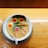 A bowl of ramen soup placed on a wooden surface. It contains various ingredients including two boiled eggs with partially soft yolks, a piece of seaweed, slices of pork, green onions, a baby corn, and a pink and white narutomaki (fish cake). A white spoon rests inside the bowl.