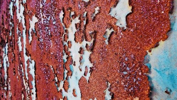 A close-up view of a rusty metal surface with peeling paint. The corrosion is unevenly distributed, revealing layers of metal and old paint with a mixture of rough and smooth textures. The rust varies in shades of brown and orange, creating an intricate pattern across the surface.