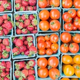 Several blue cartons filled with fresh strawberries and tomatoes are neatly arranged in a grid pattern. The strawberries appear ripe and red, with green leaves, while the tomatoes vary in color from bright red to a golden yellow, suggesting different ripeness levels or types. The image conveys the fresh produce typically found at a market or grocery store.