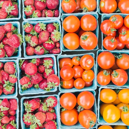 Several blue cartons filled with fresh strawberries and tomatoes are neatly arranged in a grid pattern. The strawberries appear ripe and red, with green leaves, while the tomatoes vary in color from bright red to a golden yellow, suggesting different ripeness levels or types. The image conveys the fresh produce typically found at a market or grocery store.