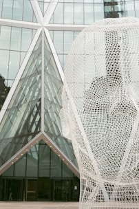 A large wireframe sculpture of a human head is positioned in front of a modern glass building with distinct geometric patterns. The structure of the building incorporates triangles, adding a sense of symmetry and complexity to the scene. The materials used are predominantly glass and metal, giving it a sleek and contemporary aesthetic.