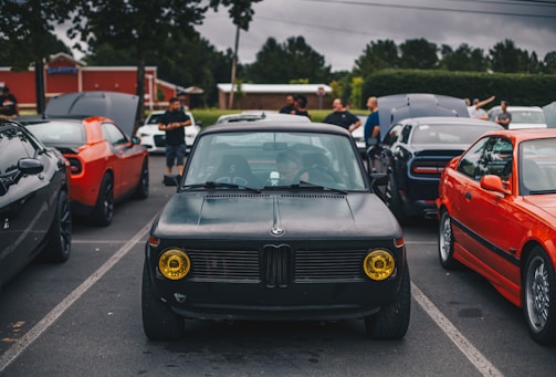 A parking lot scene with several cars of different models and colors parked closely together. The central focus is on an older black car with distinctive yellow headlights. Surrounding it are people casually standing around, some with open car hoods, as if inspecting or showcasing the vehicles. The background features green trees and buildings under a cloudy sky.