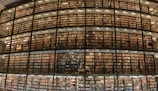 A large library with multiple stories of bookshelves filled with a vast collection of books. The shelves are arranged in a curved, floor-to-ceiling design and are accessible through a series of walkways and stairs. The lighting is warm, illuminating the books and creating a cozy atmosphere.
