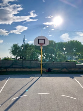 A basketball court with a single hoop in the center. The scene is set outdoors with tall green trees in the background and a clear, bright sky with scattered clouds. The sun is shining brightly, casting long shadows on the court. A net fence surrounds the area, and a backpack is placed near the base of the hoop.