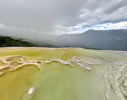 A vast, naturally formed mineral deposit with a yellowish-green hue stretches out, creating a surreal landscape. The mineral layers have formed terraces filled with shallow pools. Dense cloud cover looms overhead, partially obscuring distant mountains and a hazy horizon. Vegetation is visible at the edge of the scene, adding contrasting green tones.