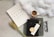 A flat lay arrangement features a white paper with the words 'Faith Hope Love' alongside a piece of wheat, a coffee cup, a black book with Asian characters on the cover, a pen, a bookmark with Asian script, a pair of glasses, and a draped piece of white fabric on a textured light gray background.