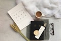 A flat lay arrangement features a white paper with the words 'Faith Hope Love' alongside a piece of wheat, a coffee cup, a black book with Asian characters on the cover, a pen, a bookmark with Asian script, a pair of glasses, and a draped piece of white fabric on a textured light gray background.