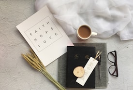A flat lay arrangement features a white paper with the words 'Faith Hope Love' alongside a piece of wheat, a coffee cup, a black book with Asian characters on the cover, a pen, a bookmark with Asian script, a pair of glasses, and a draped piece of white fabric on a textured light gray background.
