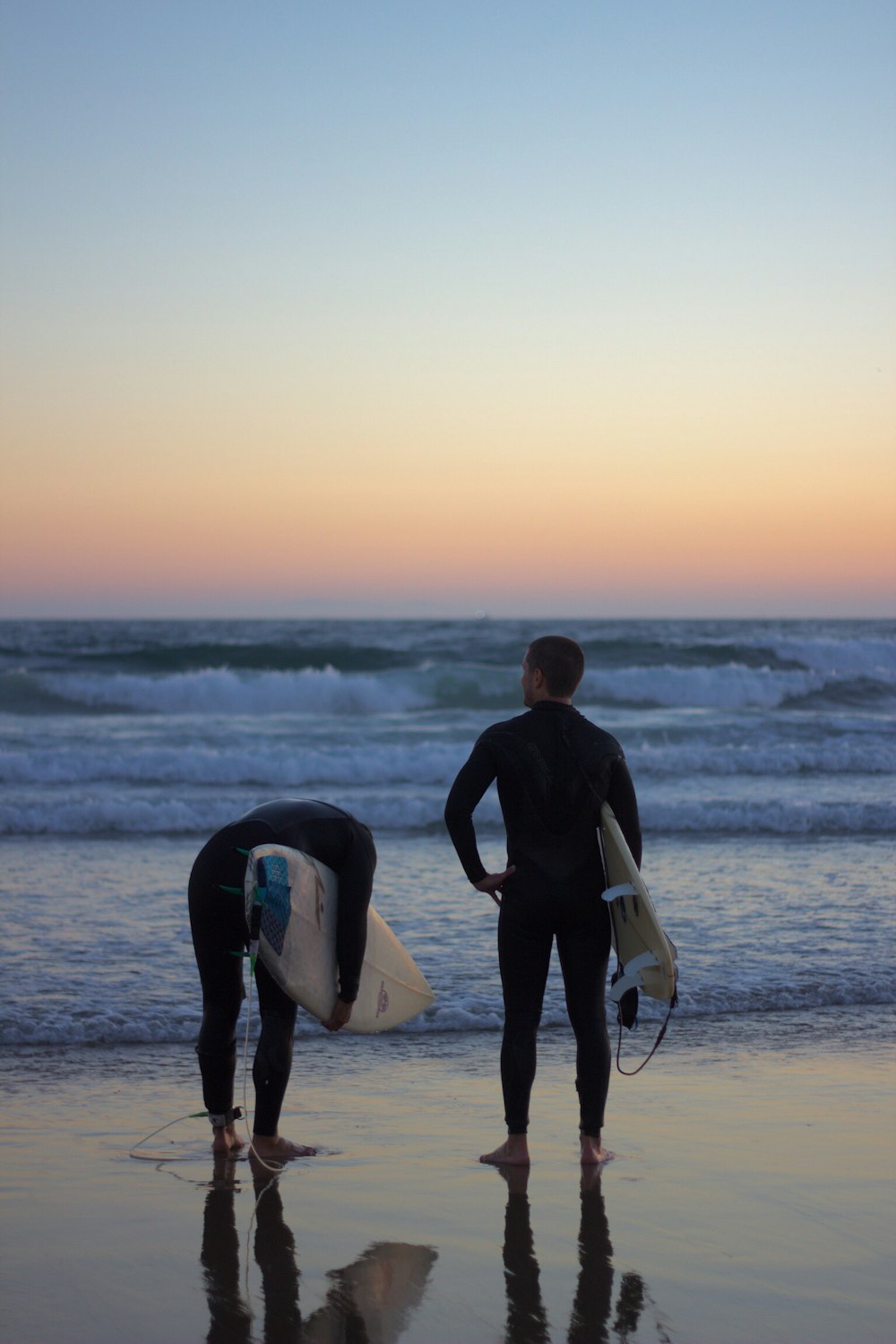 Dos personas con traje de neopreno negro sosteniendo tablas de surf de pie en la playa