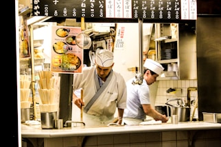 Two chefs are working in a small kitchen, possibly in a Japanese restaurant. They are wearing traditional white uniforms and hats. The kitchen is compact and organized, with signs displaying menu items and prices in Japanese above them. Various kitchen utensils, dishes, and cooking equipment are visible around them.