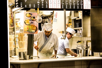 Two chefs are working in a small kitchen, possibly in a Japanese restaurant. They are wearing traditional white uniforms and hats. The kitchen is compact and organized, with signs displaying menu items and prices in Japanese above them. Various kitchen utensils, dishes, and cooking equipment are visible around them.