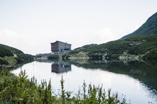 A serene mountain landscape featuring a modern building situated near a calm lake, surrounded by lush green hills. The reflective surface of the water mirrors the building and the greenery, creating a symmetrical and peaceful scene. The sky is bright with a few white clouds.