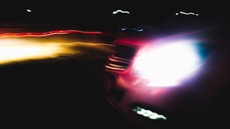 Side view of a car driving at night with bright headlights casting long beams and lens flares.