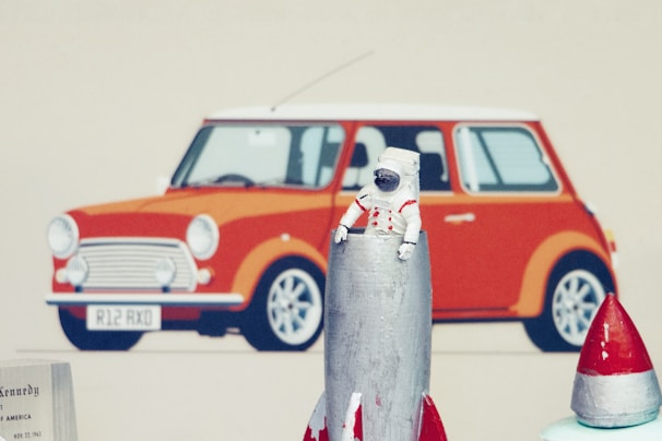 A miniature astronaut figure is positioned on top of a metallic rocket model, with a retro-style red car illustration in the background. The car is reminiscent of a classic Mini Cooper with a white roof and distinctive round headlights.