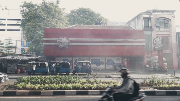 The scene captures a bustling urban street with multiple blue auto rickshaws parked alongside a red and white building with closed shutters. A blurred person on a motorcycle passes by in the foreground, while trees and various signage are visible in the background. The atmosphere is hazy with a sense of early morning or late afternoon light.