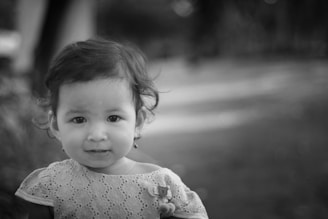 A young child with short curly hair and wearing a lace dress is looking directly at the camera. The background is softly blurred, creating a bokeh effect, which brings focus to the child's face. The image is in black and white, adding a classic and timeless feel.