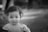 A young child with short curly hair and wearing a lace dress is looking directly at the camera. The background is softly blurred, creating a bokeh effect, which brings focus to the child's face. The image is in black and white, adding a classic and timeless feel.
