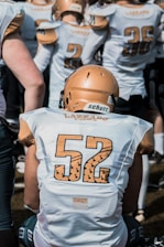 A group of football players wearing white jerseys with numbers on them. The focus is on a player with the number 52 on their back, wearing a gold helmet. Other players are visible in the background, also dressed in similar attire, with some visible numbers like 2 and 36.