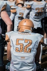 A group of football players wearing white jerseys with numbers on them. The focus is on a player with the number 52 on their back, wearing a gold helmet. Other players are visible in the background, also dressed in similar attire, with some visible numbers like 2 and 36.