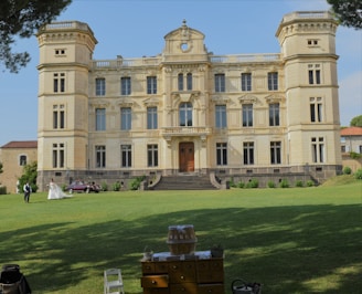 An elegant and ornate chateau-like building with multiple windows and decorative architectural elements stands on a well-kept lawn. A couple in formal attire, possibly newlyweds, are walking on the grass. A classic car is parked near the building. In the foreground, there is a small table or stand with decor items, partially shaded by surrounding trees.