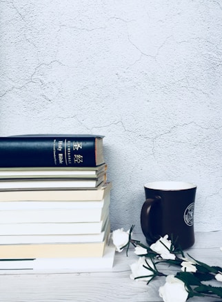 A neatly arranged stack of books sits on a wooden surface, placed against a textured gray wall. A black coffee mug is positioned next to the books. Several white flowers lie horizontally beside the mug, adding a touch of elegance.