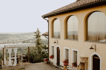 A picturesque countryside house with large arched windows and terracotta roofing. In the foreground, there is an outdoor seating area with a canopy and curtains, surrounded by lush greenery and potted red flowers. The background reveals a scenic view of rolling hills and trees on a clear day.