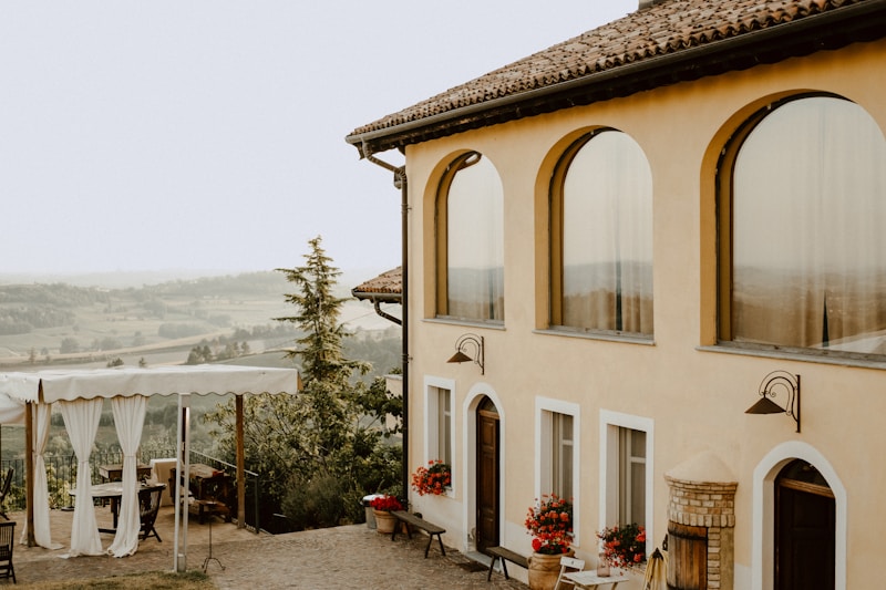 A picturesque countryside house with large arched windows and terracotta roofing. In the foreground, there is an outdoor seating area with a canopy and curtains, surrounded by lush greenery and potted red flowers. The background reveals a scenic view of rolling hills and trees on a clear day.