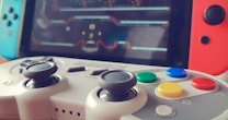 Close-up view of a video game controller with colorful buttons including red, yellow, green, and blue. A gaming console screen in the background, slightly blurred, displays a game interface. The controller features dual analog sticks and buttons labeled with letters and symbols.