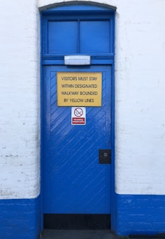 A blue door set into a white brick wall features two signs. The larger sign, with a yellow background, instructs visitors to stay within designated walkways bounded by yellow lines. The smaller, red sign highlights that children must be closely supervised. The door has diagonal wooden panels and a metal lock.