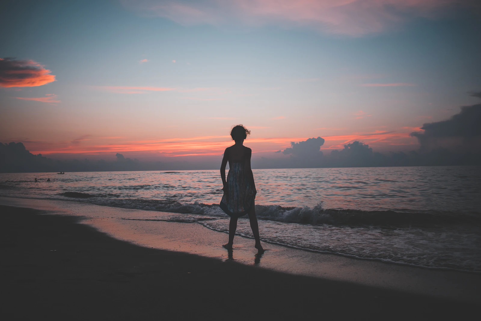 A woman standing on the seashore during golden hour, warm back-lit sunlight on the horizon