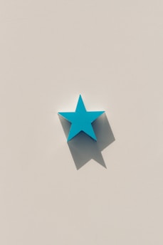 A stylized three-dimensional turquoise star sits against a beige background. The star casts a distinct shadow, emphasizing its shape and giving the image a minimalist aesthetic.