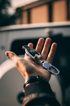 A hand holds a car key fob with shiny metallic parts, set against a blurred outdoor background, suggesting a vehicle in the surroundings.