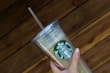 A hand holding a clear plastic tumbler with a green and white logo featuring a siren. The tumbler has a lid and straw, and it is held against a wooden, textured background.