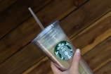 A hand holding a clear plastic tumbler with a green and white logo featuring a siren. The tumbler has a lid and straw, and it is held against a wooden, textured background.