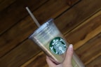 A hand holding a clear plastic tumbler with a green and white logo featuring a siren. The tumbler has a lid and straw, and it is held against a wooden, textured background.